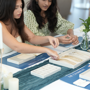 Two women examining jewelry at a table for their Charms Jewelry making workshop in Dubai for EID 2026 and there is a vase in the background.